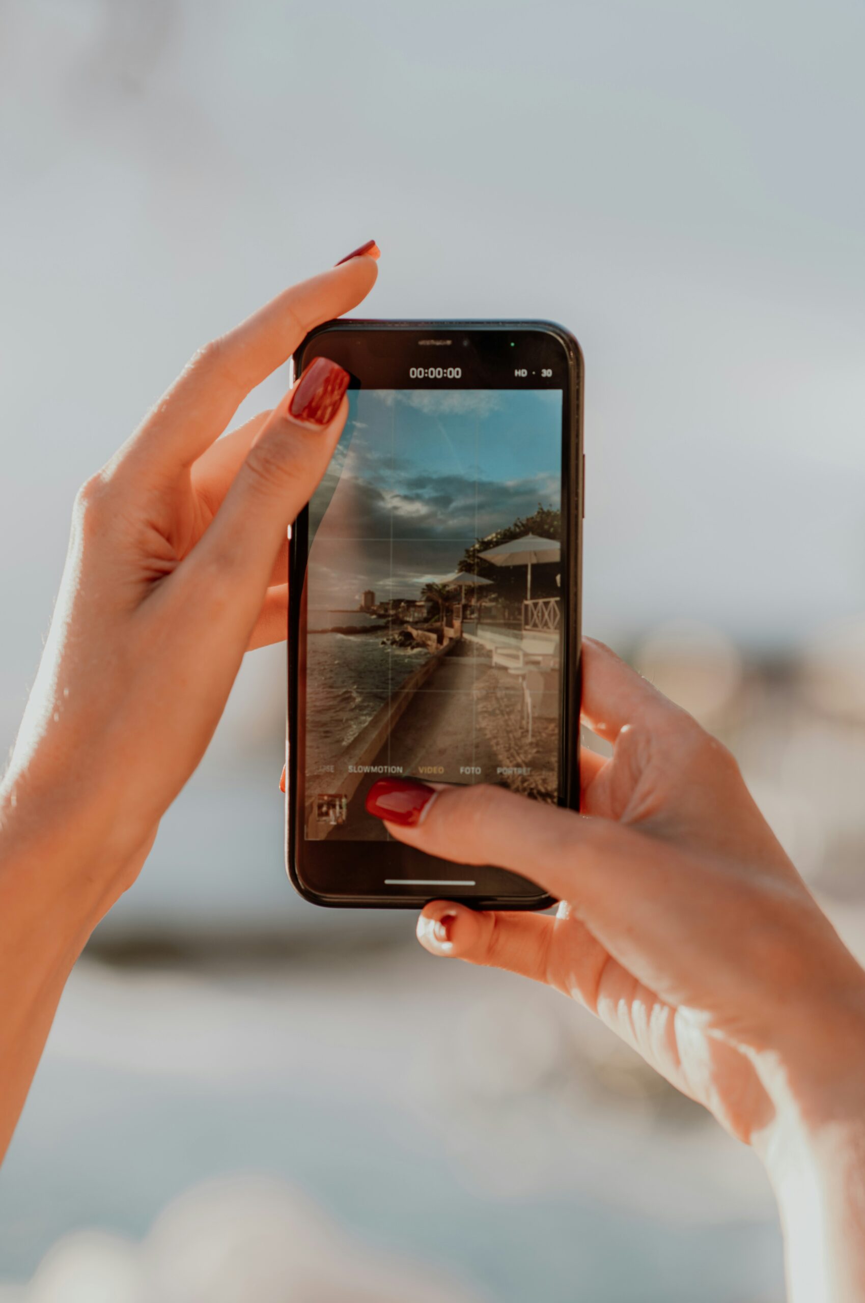 A person's hands holding a smartphone taking a video of a scenic beachside view displayed on the phone's screen, showcasing a clear sky and a serene coastal environment.